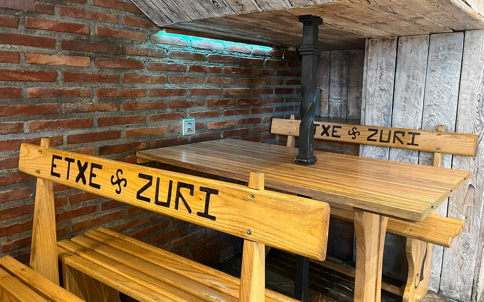 'Etxe Zuri' eta lauburua tailatuta dituzten zurezko mahaia eta bankuak, adreiluzko hormaren aurrean